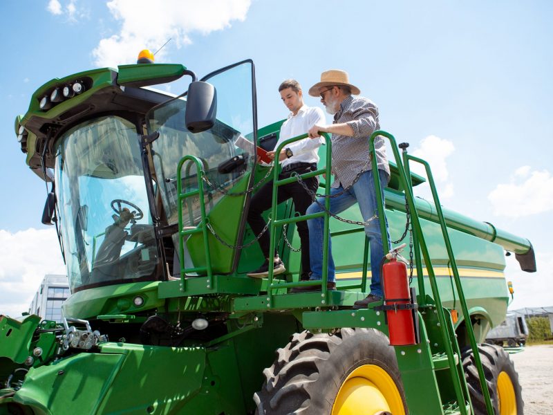Professional farmer with a modern tractor, combine at a field in sunlight at work. Confident, bright summer colors. Agriculture, exhibition, machinery, plant production. Senior man on his machine with investor.